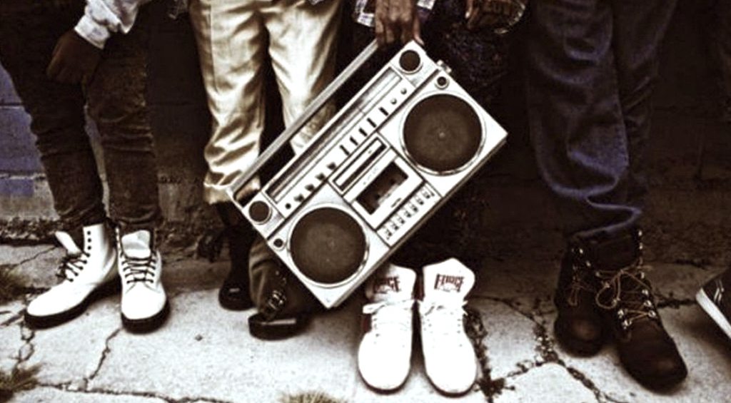 Picture of people's lower legs with differnt footwear and a large boombox in the middle
