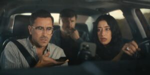 A young man with glasses sits in the passenger seat of a car looking at his phone, while a dark-haired young woman drives and looks over at his screen. A man sits in the back, in the middle seat, and his face is blurred.