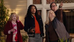 Four people (from left, a blonde older white woman, a young beautiful Black woman, a sixty-something white woman, and a bald white man in glasses and a beanie) stand in front of a Colonial house looking upwards. The third woman is pointing at something high up.