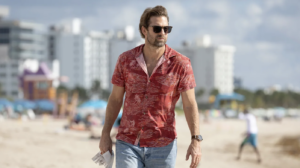 A man in a red Hawaiian shirt strolls down a beach towards the camera